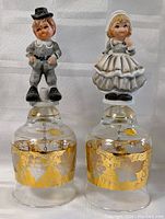 Glass bells set with gold floral decoration and boy and girl Hummel figurines on top shown front view.