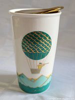 Photo showing the full side of the white ceramic travel mug featuring a turquoise and gold hot air balloon with mountain and water scenery at the base.
