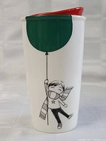 Front and side view of white ceramic Starbucks travel mug with red lid and cartoon design