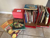 Wide view showing multiple vintage cookbooks arranged beside a red Betty Crocker recipe box on a tile floor.