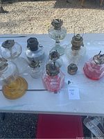 Overview of all eight hurricane lamps arranged on a white table outdoors, showing various sizes, colors and glass shapes.