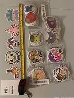 Photo showing multiple small bundles of stickers lined up, including axolotl designs, fruit and donut stickers, and some animal and character stickers. A tape measure provides size reference showing stickers around 2-3 inches.
