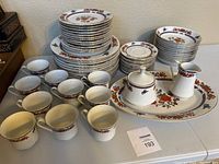 Stack of dinner plates, side plates, bowls, coffee cups, sugar bowl with lid, creamer, and large serving platter displayed on a table.