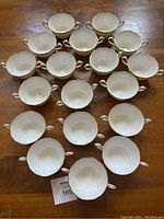Top-down view of 18 cream-colored double handled soup or dessert cups arranged in rows on a wooden table.