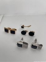 Set of six pairs of cuff links on white background, showing gold plated rectangular and oval black onyx stones and silver plated rectangular black onyx stones with textured borders.