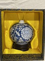 The porcelain vase is shown inside the decorative box with yellow satin lining, placed on the wooden base. The vase has blue floral patterns against a white background.