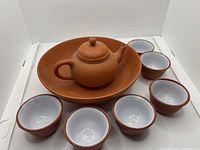 Small terracotta teapot with lid placed inside a large matching tray along with six small tea cups arranged around it