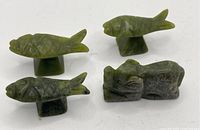 Photo shows three green jade figurines: two fish mounted on stands and one pig. Detail of carving on fish scales and pig facial features visible.