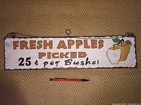 Photo of rectangular metal sign with scalloped edges. White background with orange text: "FRESH APPLES PICKED 25¢ per Bushel", red and yellow apple illustrations on right, attached wire hanger.