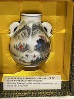 Porcelain rosy bowl bottle shaped pen holder nestled in yellow silk lining inside a black decorative silk box, showing its full front with landscape and human figure artwork.