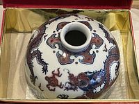 Top view of the small round white porcelain vase with intricate red and blue dragon design, placed inside a red and gold decorative box.