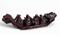 Side view of the dragon boat boxwood carving showing the figures of the Eight Immortals arranged onboard beneath the dragon head at the front and curled tail at the back, dark polished wood finish.