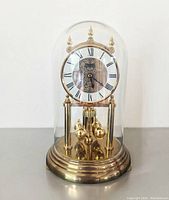 Front view of clock under glass dome with ornate dial and rotating pendulum balls on brass base.