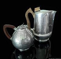 Two-piece set showing a hammered pewter creamer with rounded body on left and a polished aluminum Picquot Ware teapot with angular wooden handle on right, both placed on reflective black surface.