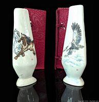 Two slender white eggshell porcelain vases standing next to their original red fabric boxes, one vase featuring a galloping horse and the other an eagle in flight