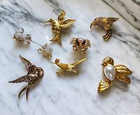 Photo showing seven gold-tone figural pins arranged on a marble surface, including hummingbird, crab, mice, and bird designs.