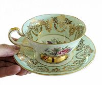 Side view of mint green teacup with gold scrollwork and central fruit and floral motif, held by hand to show details