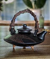 Miniature Satsuma Japanese stoneware teapot with dark earthy body, tripod feet, short spout, and woven handle.