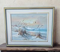 Frontal view of the framed original seaside painting resting on a wooden surface against a beige wall.