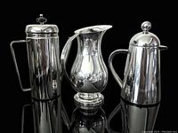 Front view of the three serveware items on black reflective surface showing French press, copper jug, and stainless carafe.