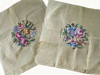 Two square needlepoint canvases side by side showing floral needlepoint designs on neutral background fabric.