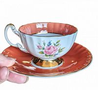 Hand holding vintage Aynsley teacup with scalloped oban shape and orange exterior. The cup is white inside with a pink rose and blue flower floral pattern on the side, gold trimmed rim and handle. The matching orange saucer has a pink floral center and gold rim.