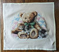 Full view of completed cross stitch artwork showing a teddy bear with a green bow surrounded by three kittens seated on a pastel patchwork cushion on beige fabric.