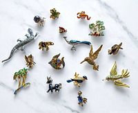 Top-down view of complete lot showing all 19 vintage figural pins arranged on white marble surface, including various birds, insects, palm trees, animals, a marcasite lizard and fish pins.