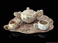 Front view of the soapstone teapot with dragon carvings, three teacups, and tray with inlay on black background