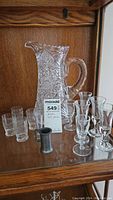 Full view of cut glass pitcher surrounded by vintage short shot glasses and taller cordial glasses, all displayed on wooden shelf