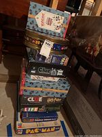 Stack of varied board games and puzzle boxes including Pac-Man, Clue, Life, Mickey Mouse Game, and a 1000-piece jigsaw puzzle, photographed on floor.