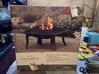 Box front showing the Threshold steel fire bowl fire pit with image of fire burning and product details including powder-coated finish and folding stand.