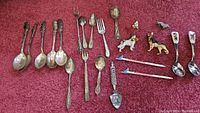 Various flatware pieces laid out on red carpet including spoons with ornate handles, cartoon character spoons, key-shaped flatware, and airplane-shaped stirrer sticks.