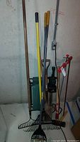 Garden tools arranged standing against wall including multiple rakes, a shovel, trimmer, and manual tiller.