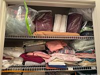 Two shelves of assorted folded linens including sheets, pillowcases, blankets, and towels in various colors and patterns stored on wire shelving.