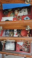 View of multiple drawers showing various small costume jewelry items including pins, bracelets, necklaces, loose rhinestones, and faux pearl earrings.