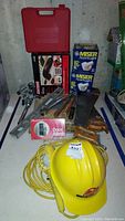 Photo showing assorted tools and electrical items including a yellow Bechtel hard hat, corded cage light, door alarm packaging, hand tools, saw blades, shelving brackets, and boxes of Miser light bulbs.