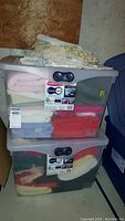 Two clear plastic storage containers filled with assorted folded towels and linens in various colors (pink, white, red, cream, gray). Some towels are fuzzy terry cloth.