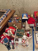 Overview of Christmas decorations including boxed ornaments, stockings, Santa figurines, and musical snow globe placed on floor.