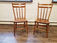 Front view of two antique wooden chairs showing contoured seats and vertical slat backs on a wooden floor.