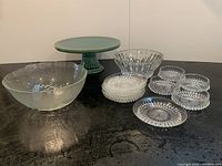 Photo showing green ceramic cake pedestal, large glass bowl, medium bowl, six plates, and four smaller bowls arranged on black surface.