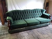 Frontal angled view showing the green velour upholstery, wooden carved armrests, three seat cushions, and tufted backrest of the sofa on a tiled floor.
