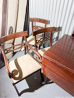 Photo of three wooden dining chairs with intricate back designs, light beige upholstered seats, and part of the wooden dining table visible in the corner. Shows condition and style of chairs.