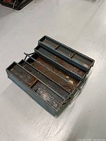 Open view of the antique metal toolbox showing three tiered fold-out compartments with rust and worn paint.