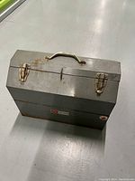 Closed antique gray metal toolbox with gold-tone handle and two latches, showing some surface rust and wear.