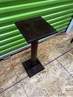 Front angled view of the dark wooden column plant stand on a plywood floor against a green metal background.