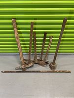 Front and side angle showing all heavy hand tools including 3 pick axes, 2 axes, tamping tool, and pry bar against a green corrugated background.