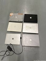 Six laptops consisting of five Apple MacBooks and one Microsoft Surface laid flat on a gray floor. Visible scratches, stickers, and wear marks on MacBooks. Surface laptop is silver with attached charger.