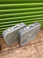 Two box fans shown side by side, positioned on a plywood floor with a green metal background. Fans are rectangular and plastic, one with a rotary knob and handle, the other with push buttons and a wrapped cord.