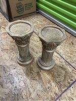 Pair of decorative wood plant stands with classic fluted columns and ornate detailing, octagonal top surface, photographed on wood floor with surrounding storage items.